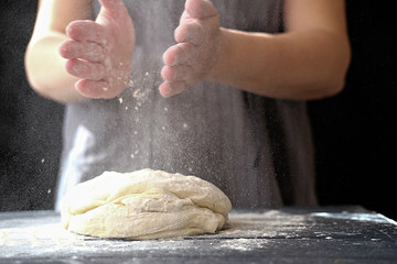 Fototapeta premium Making dough by hands at bakery or at home. Flour cloud in the air. Close up of woman chef hands preparing for kneading the dough on the table, powdering with flour. dough for bread, pasta or pizza