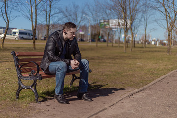 Fototapeta na wymiar A young adult man in a black leather jacket and jeans sits on a bench in a park on a city street on a sunny day