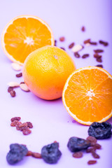 Oranges whole and cut in half on a purple background. Garnished with raisins, seeds, cashews, hazelnuts.