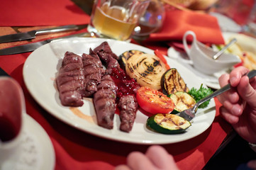 Grilled beef served with grilled vegetables, dish plate on а red tablecloth, fork, hands, close-up view with blurred background. Restaurant food, delicious dish.