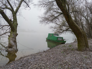An old boat moored along the river bank in a willow grove. Dense fog in the river valley and frozen nature in a morning. A gloomy autumn landscape of frozen nature in a cold and silent fog.