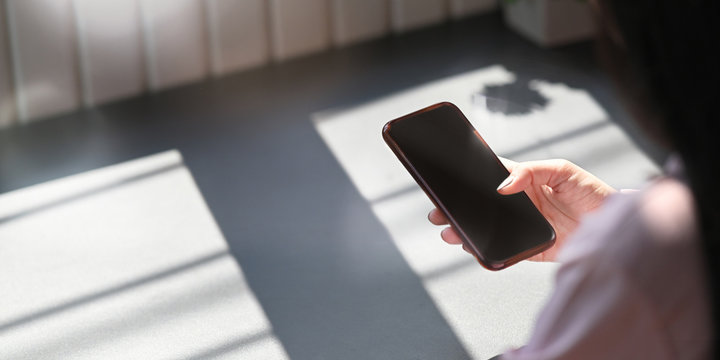 Cropped image of beautiful woman hands holding and using a blank screen smartphone while sitting at the working desk over comfortable working room as background.