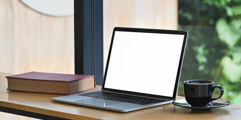 Photo of computer laptop with white blank screen, book and coffee cup putting on wooden counter bar over comfortable living room glass wall as background.
