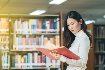 Asian young student in white casual suit reading  the book in library of university or colleage.Standing and reading on a wooden table and bookcase in the background.Back to school