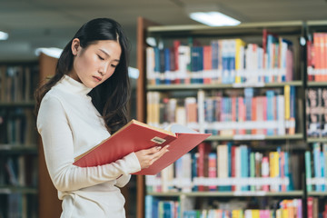 Asian young student in white casual suit reading  the book in library of university or colleage.Standing and reading on a wooden table and bookcase in the background.Back to school