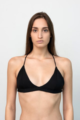 Fototapeta premium Model snaps. Young beautiful model with dark hair in black underwear or swimwear looking at camera while standing isolated over white background. Vertical shot. Front view