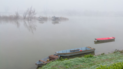 Moored old rusty fishing row boats on water's edge of calm river and fisherman's huts in front of river island deep in the fog. Gloomy autumn landscape wrapped in a thick fog.