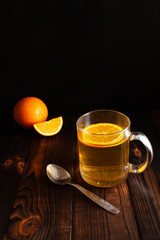 Tea with orange, metal spoon, sliced ​​orange, wooden brown background. Citrus drink in a transparent cup, empty space for text. Food composition on a black background in low key.