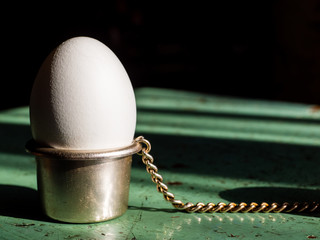 A white egg stands in a metal egg stand on the table, on a black background, close- up, copy space....