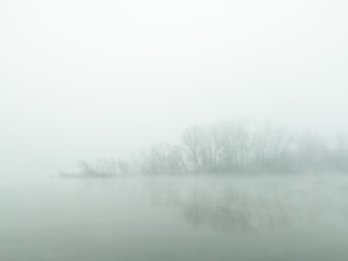 Obraz na płótnie Canvas Mysterious and ghostly river island obscured by dense fog which float above flat water surface and slips through between trees. The gloomy silent autumn landscape of the river island in the thick fog.