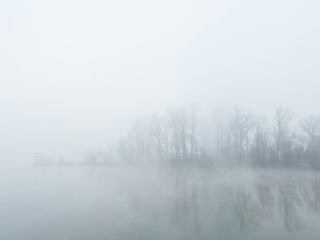 Fototapeta na wymiar Peaceful autumn scene of mystical river island with a deciduous grove in deep fog early in morning. Calm and silent nature scene covered with thick fog.