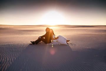 Fototapeta na wymiar The bride and groom are sitting in the desert on the sand, a beautiful sunset, lovers man and woman, a new family, love and tenderness, a beautiful couple in love, designer wedding dress, wedding day