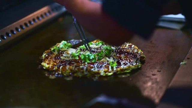 Okonomiyaki, Japanese pizza: closeup of Asian chef hands cutting Okonomi-yaki - cabbage pancake version of Potato Latkes (Traditional Jewish Hanukkah food) on professional kitchen grill background