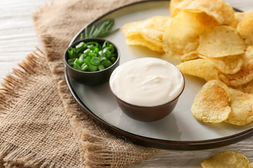 Plate with tasty sour cream, green onion and potato chips on table