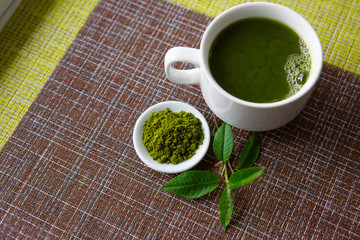 Japanese matcha green tea is poured into a white mug and on a white saucer in powder. Tea set on a textured napkin of natural flowers, decorated with a branch of green leaves. Background with space fo