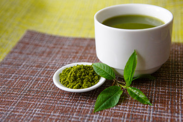 Japanese matcha green tea is poured into a white mug and on a white saucer in powder. Tea set on a textured napkin of natural flowers, decorated with a branch of green leaves. Background with space fo