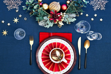 Christmas table setup with dark red white plates, red paper ring and poinsettia, golden utensils. Red, green and golden gilded decorations. Flat lay, top view on dark green linen textile table cloth.