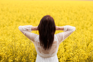 Young woman in a white dress raised her hands above a head in a background of the yellow field with rape flowers. Concept of wedding photo in spring with copy space.