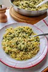 Orzo primavera with green veggies in vintage place over white marble table. One pot pasta with serving spoon, pepper grinder and napkin on set table. 