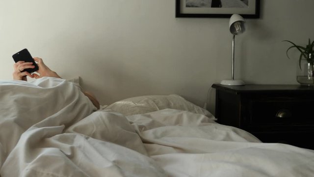 Woman stretching in bed reaching for her phone after waking up in the morning