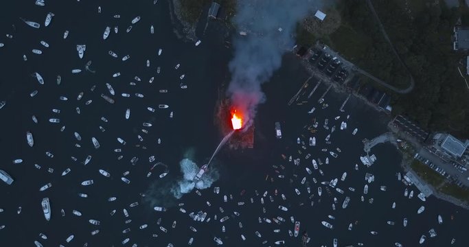 Unique, high altitude, top down, turning aerial drone footage of small motor boats and sail boats gathering around and witnessing the biggest bonfire event on Earth at Alesund, Norway at dusk.