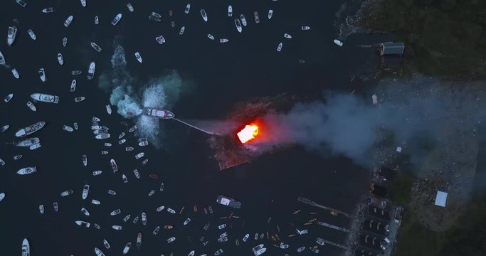 Unique, high altitude, top down, turning drone footage of small motor boats and sail boats gathering around and witnessing the biggest bonfire event on Earth at Alesund, Norway at dusk.
