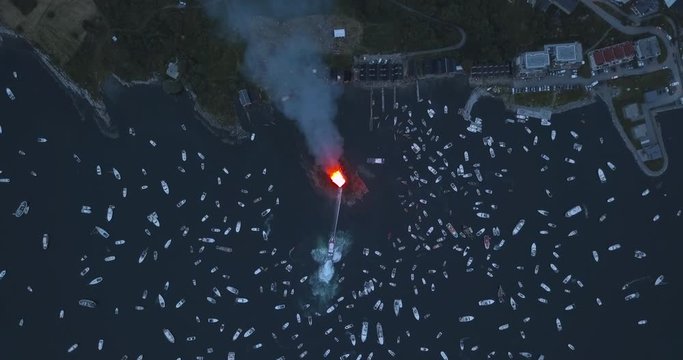 Extremely unique, high altitude, top down, turning aerial footage of small motor boats and sail boats gathering around and witnessing the biggest bonfire event on Earth at Alesund, Norway at dusk.