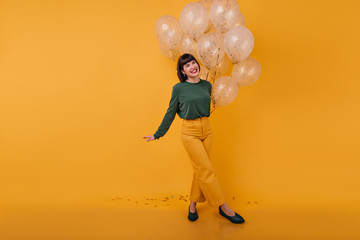 Fototapeta na wymiar Full-length portrait of laughing woman standing with legs crossed on yellow background. Indoor shot of romantic birthday girl dancing with golden balloons.