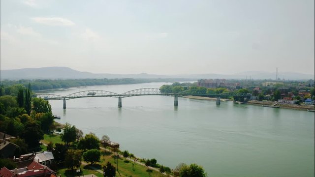 M&aacute;ria Val&eacute;ria bridge between Hungary and Slovakia. A cargo ship floating up in the river Danube under a busy bridge. Another ship turn around in the distance. People walking in a park near river.