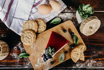 Slice of blackberry cheesecake on a wood counter, with some berries, biscuits, eggs, green leaves, and red lines on the side.