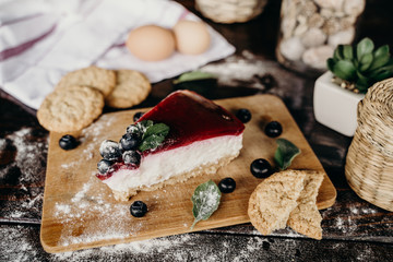 Slice of blueberry cheesecake on a wood counter, with some berries, biscuits, eggs, green leaves, and red lines on the side.