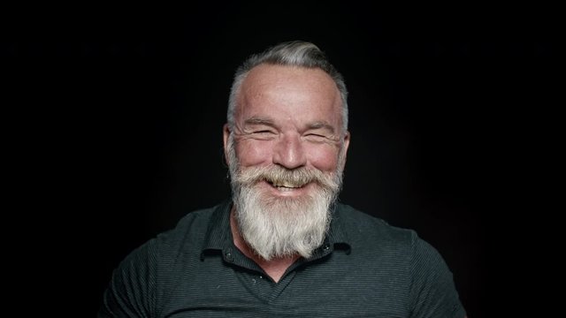 Close up of cheerful senior man with a white beard. Retired old man laughing on black background.

