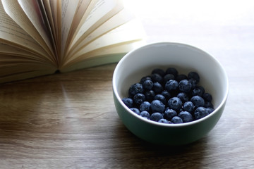 Fototapeta na wymiar Bowl of fresh blueberries and open book on a table. Selective focus.
