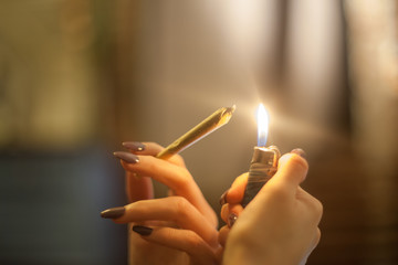 Young woman with pretty manicured nails is holding a lighter and about to spark and light a legal pre rolled marijuana bud joint in organic hemp rolling papers for THC smoking - obrazy, fototapety, plakaty