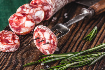 Dried sausage on a rustic board with rosemary and spices. A slice of sausage on a fork. German meat products. Close-up.