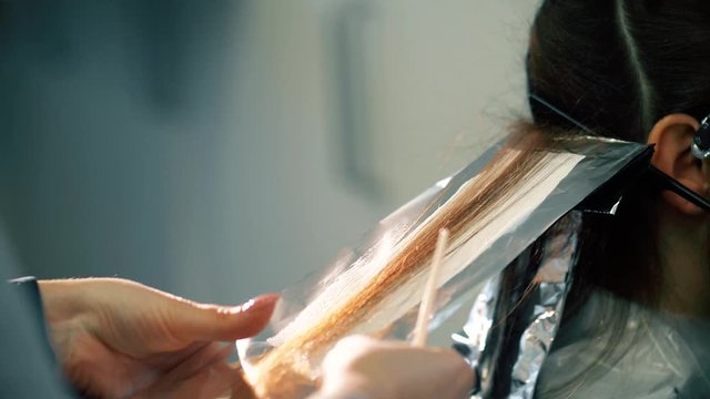Beauty spa client having color of hair changed. Colorist lays hair of woman on foil with hair coloring paste to prepare for wrapping. Close up view of hands of stylist and hair technique in salon.