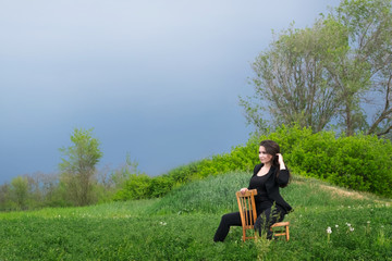 Naklejka na ściany i meble The girl in black sits on a wooden chair with her back in a green field on nature. Blue sky and place to insert.