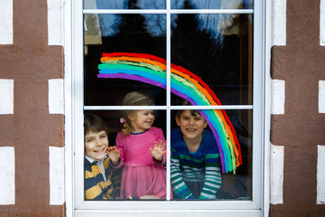 Three kids, two school kids boys and toddler girl with rainbow painted with colorful window color during pandemic coronavirus quarantine. Children painting rainbows with the words Let's all be well.