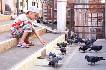 A little girl feeds pigeons. The girl is sitting on the stairs. The girl in the cap. A child in Venice. Birds eat the bread. To share food. Love nature. Help the weak. One in the city.