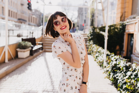 Glad young lady in white dress smiling during outdoor summer photoshoot. Short-haired brunette girl in sunglasses ejoying spring.
