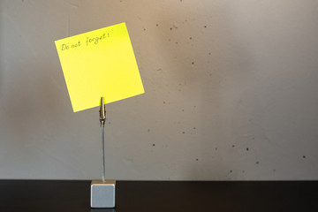The inscription "Do not forget" on a yellow piece of paper attached to a clip standing on a black surface against a gray concrete wall.