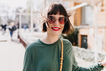 Obraz premium Laughing joyful woman in brown sunglasses posing on street background. Outdoor shot of pleased european lady in green sweater.