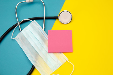 Stethoscope and pink sticky note for text above face mask on colorful background.
