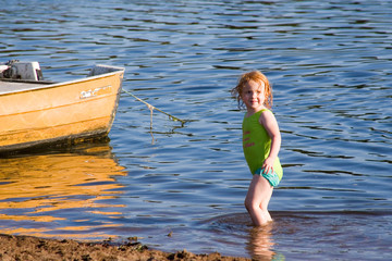 Rothaariges kleines Mädchen im grünen Badeanzug steht im Meer neben einem gelben Boot