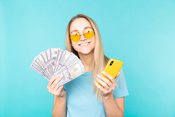 Image of young smiling woman holding yellow smartphone and banknotes in hands isolated over blue background.