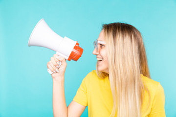 Young woman in yellow t-shirt over blue wall shouting through a megaphone and smilling.