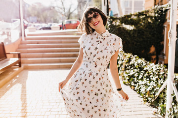 Refined girl with red lips smiling during walk in summer day. Brown-haired woman in long white...