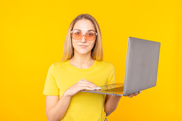 Young woman in sunglasses showing laptop computer isolated over yellow background.