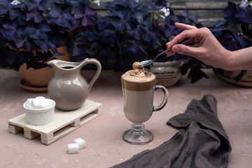 A popular Korean drink during self-isolation is Dalgona coffee. A delicious modern cold beverage on the table. Female hand hold a little spoon over a glass. Modern vertical still life.