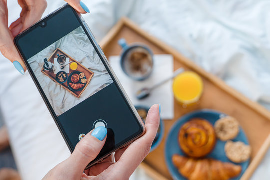 Woman using smartphone to take photos of her breakfast served in bed on wooden tray with coffee and croissants